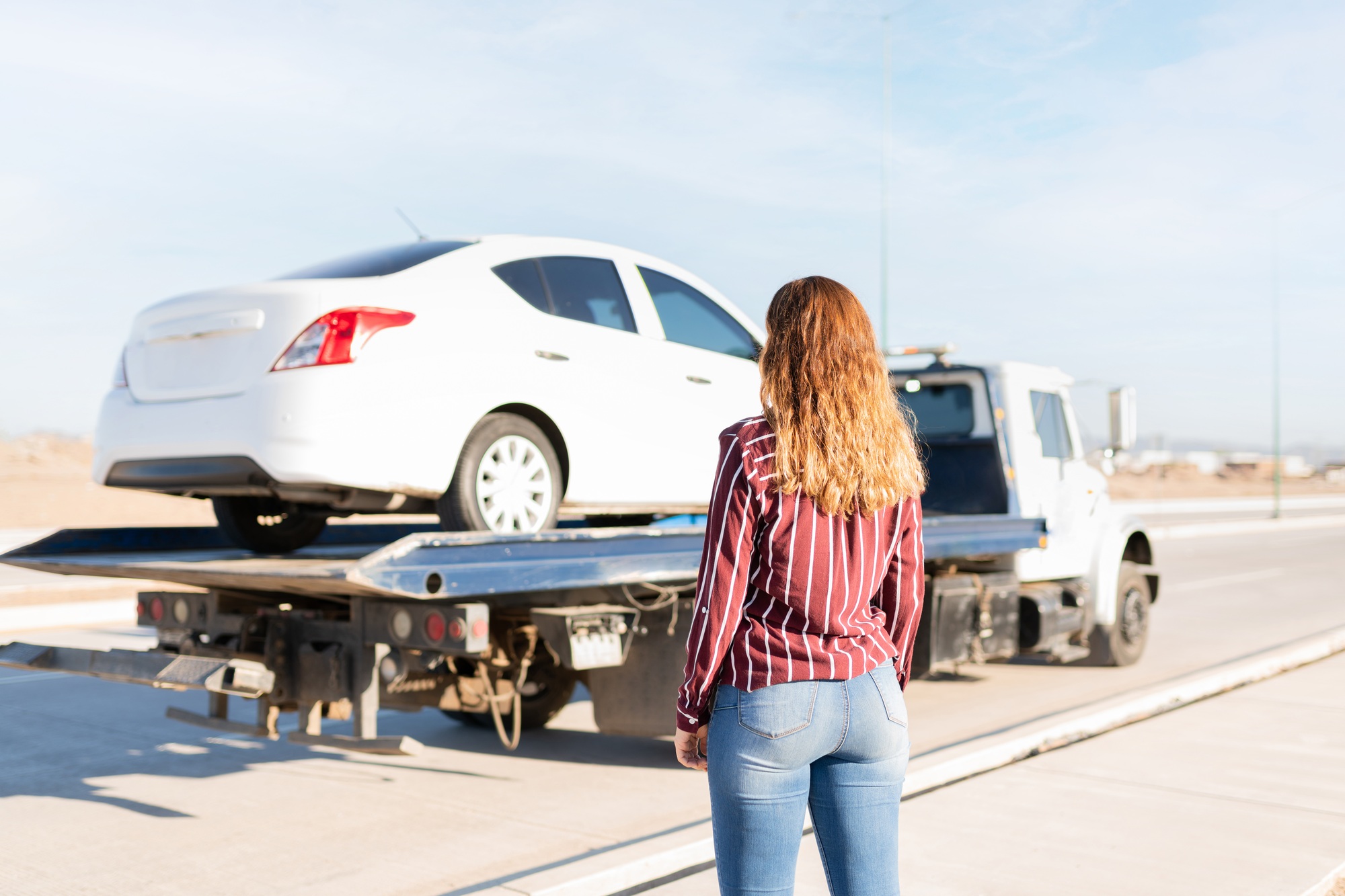 Young woman seen from behind staring at her car in a tow truck